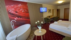 A hotel room with a large bathtub, a bed with white linens, a small round table with a vase of flowers, a desk with two chairs, a television on the wall, and a red carpet. - Kräuterhotel Heidejäger (Rotenburg)