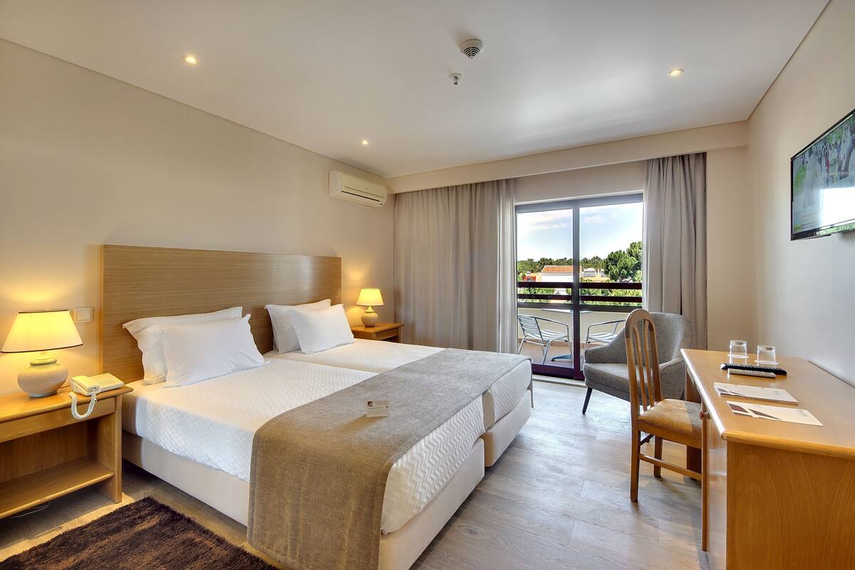 Een hotelkamer met twee bedden, een bureau, een stoel, een balkon en een televisie.