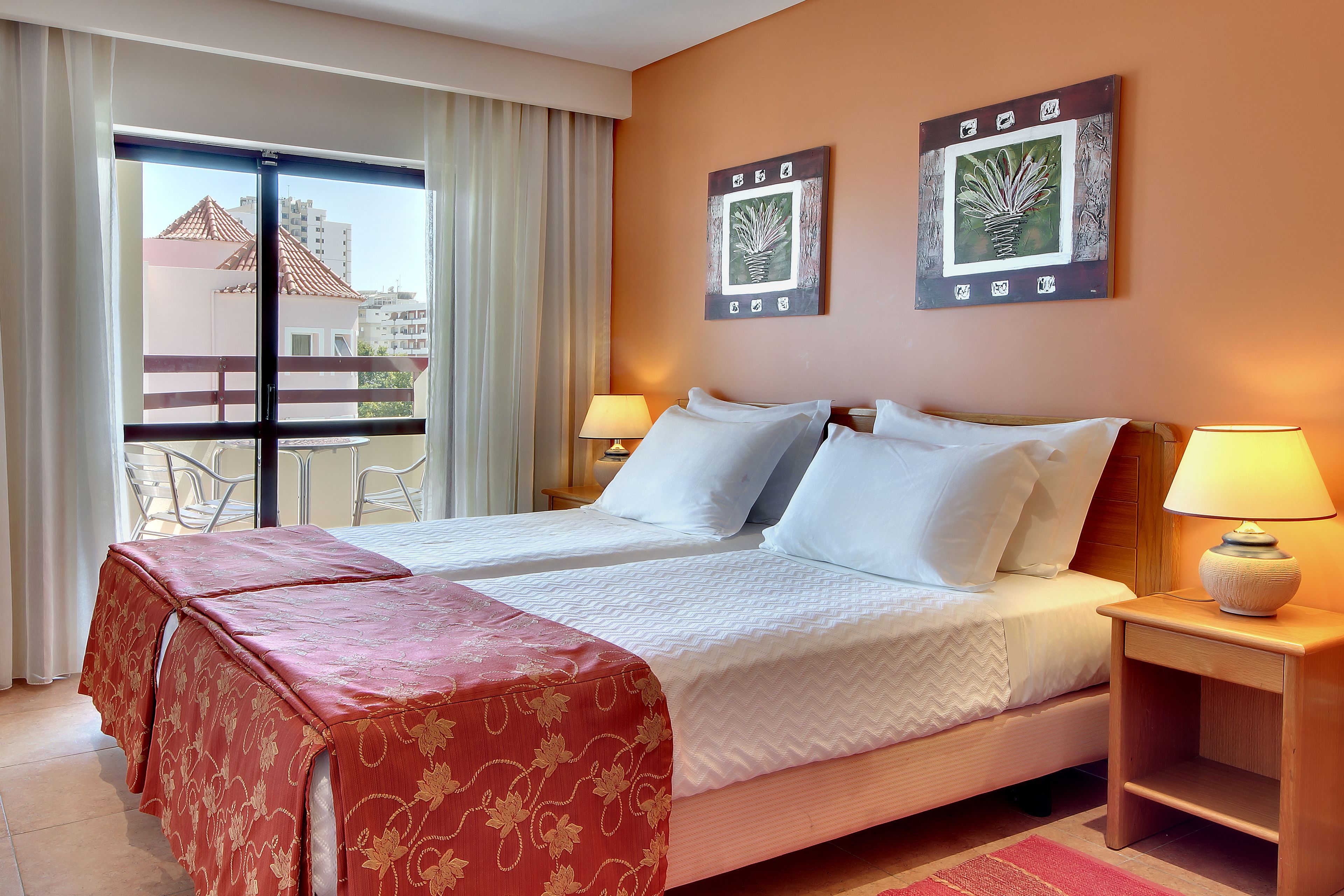 Habitación de hotel con una cama grande, dos mesitas de noche, un balcón con vista a edificios y dos cuadros en la pared.