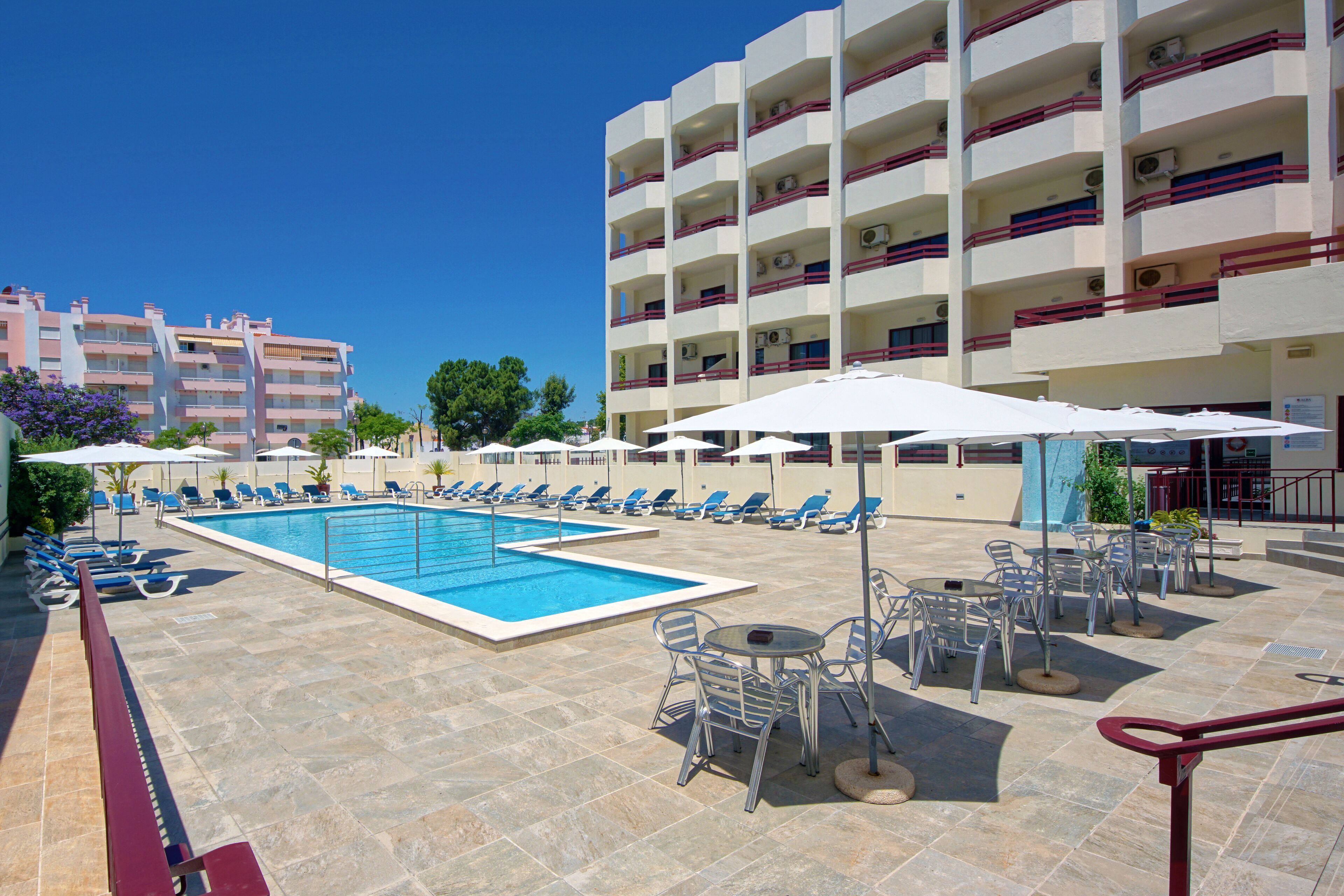 A hotel pool area with multiple lounge chairs and umbrellas, a pool, and a building with balconies.