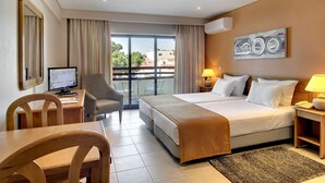 Habitación de hotel con cama, escritorio con computadora, silla, balcón con vista a edificios y un cuadro en la pared.