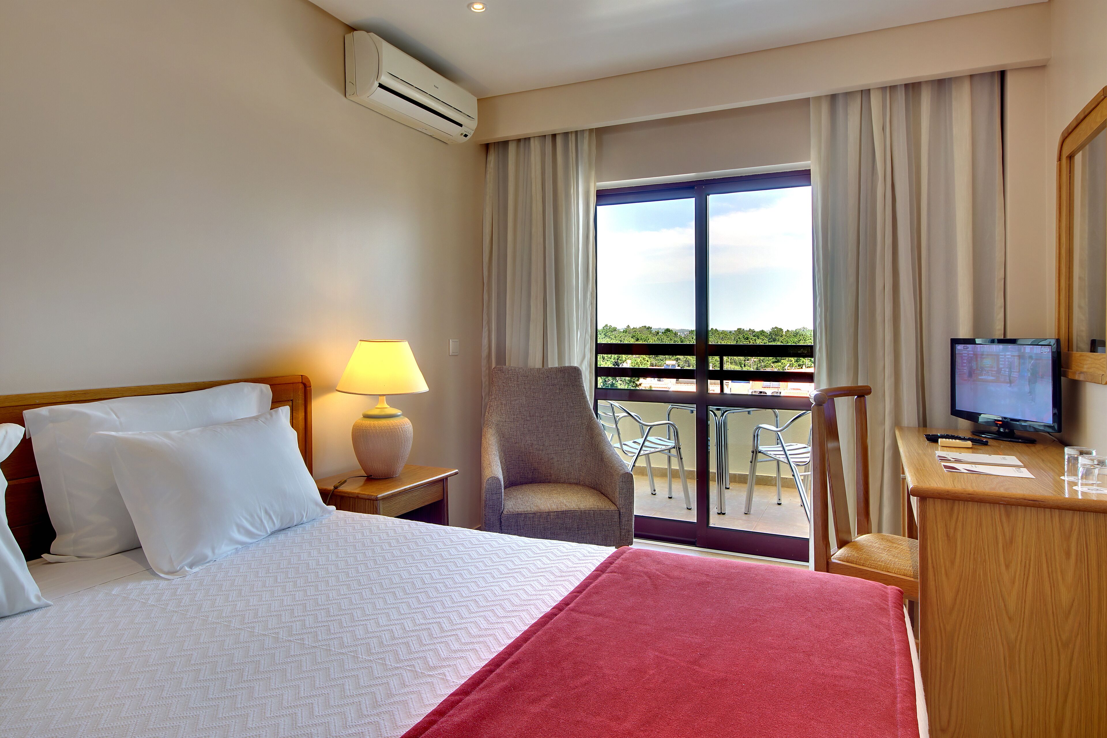A hotel room with a bed, a desk with a computer, a chair, and a balcony with a view.