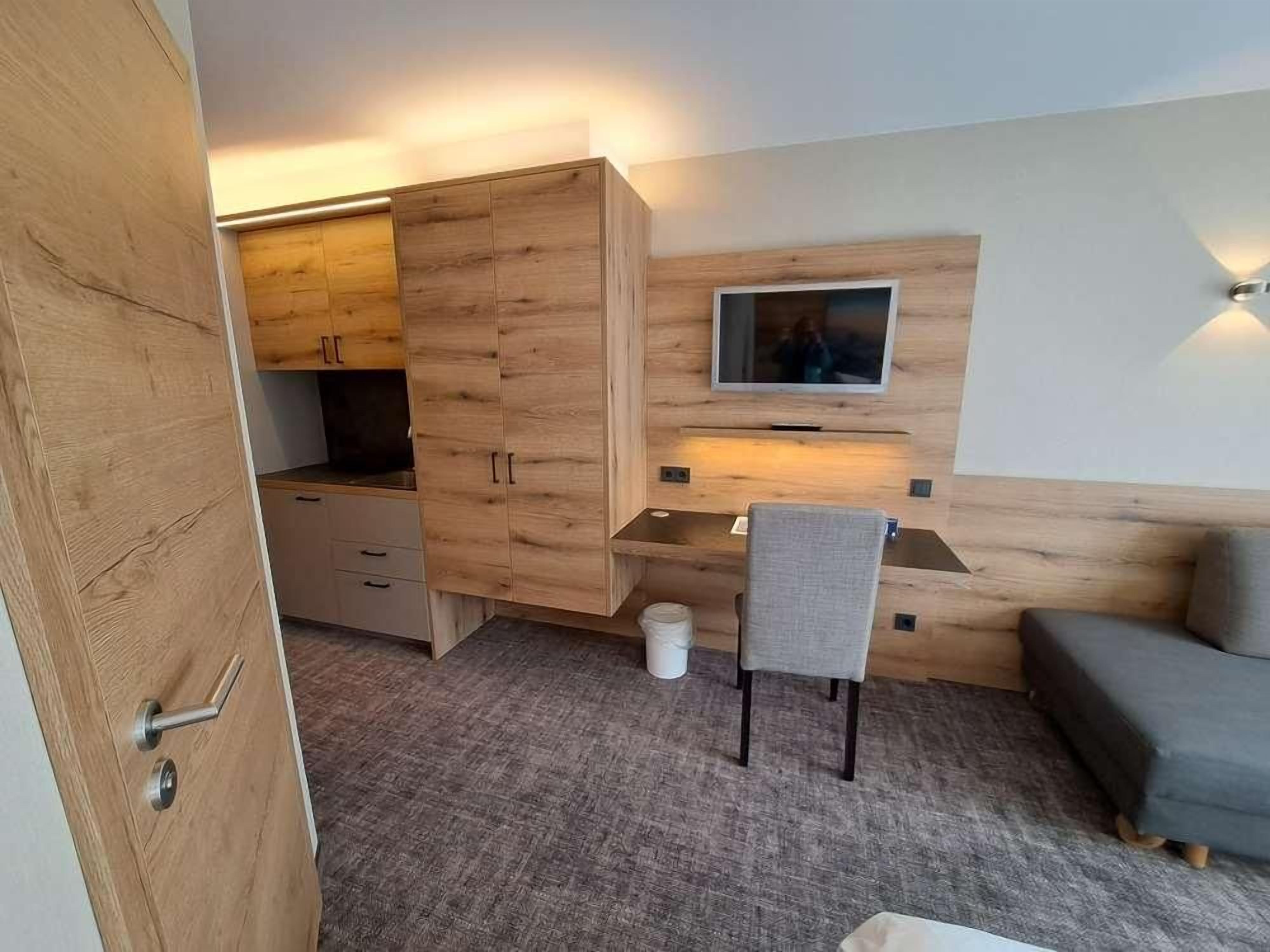 Ein gemütlicher Raum mit einem Holzschrank, einem Schreibtisch mit Stuhl, einem an der Wand befestigten Fernseher und einer Couch.