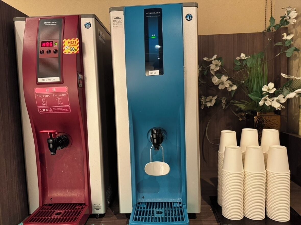 Dos dispensadores de agua, uno rojo y otro azul, con pilas de vasos desechables a un lado.