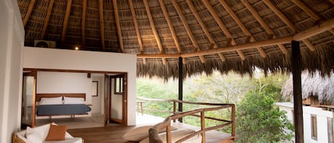 A room with a thatched roof, wooden flooring, a bed, a wicker sofa, and a view of greenery outside.