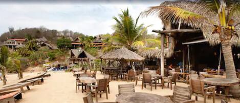 Ein Restaurant direkt am Strand mit Holztischen und -stühlen, Reetdachschirmen und Blick auf den Strand und die umliegenden Gebäude.