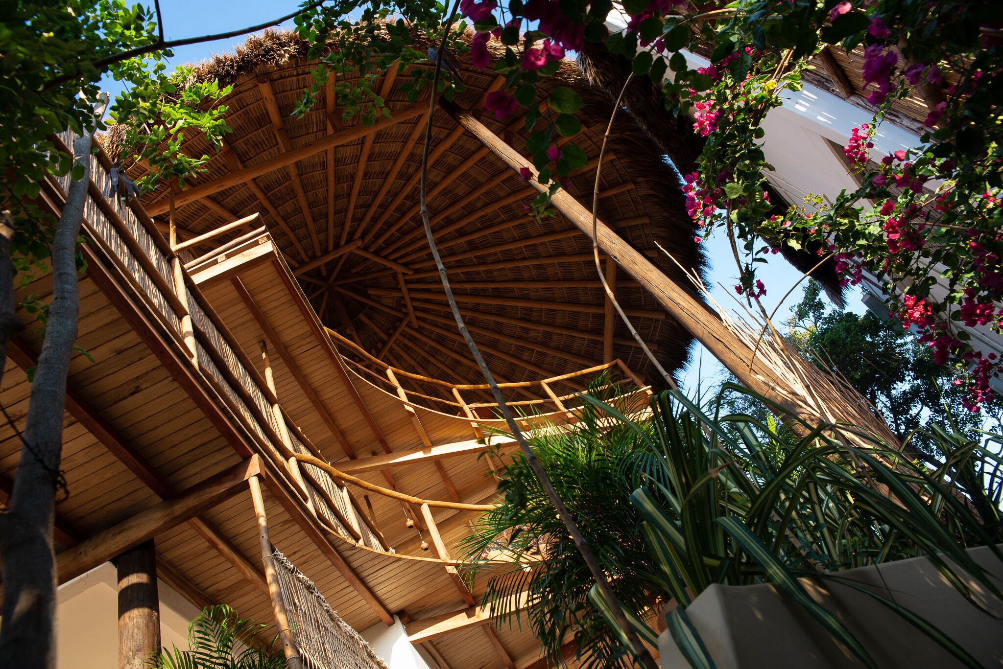 Une structure en bois à plusieurs niveaux, surmontée d’un toit de chaume, et entourée d’une végétation luxuriante et de plantes en fleurs.