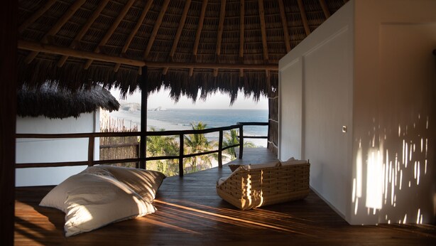 A room with a thatched roof, wooden flooring, a white door, and a view of the ocean. - El Alquimista Yoga & Spa (San Pedro Pochutla)