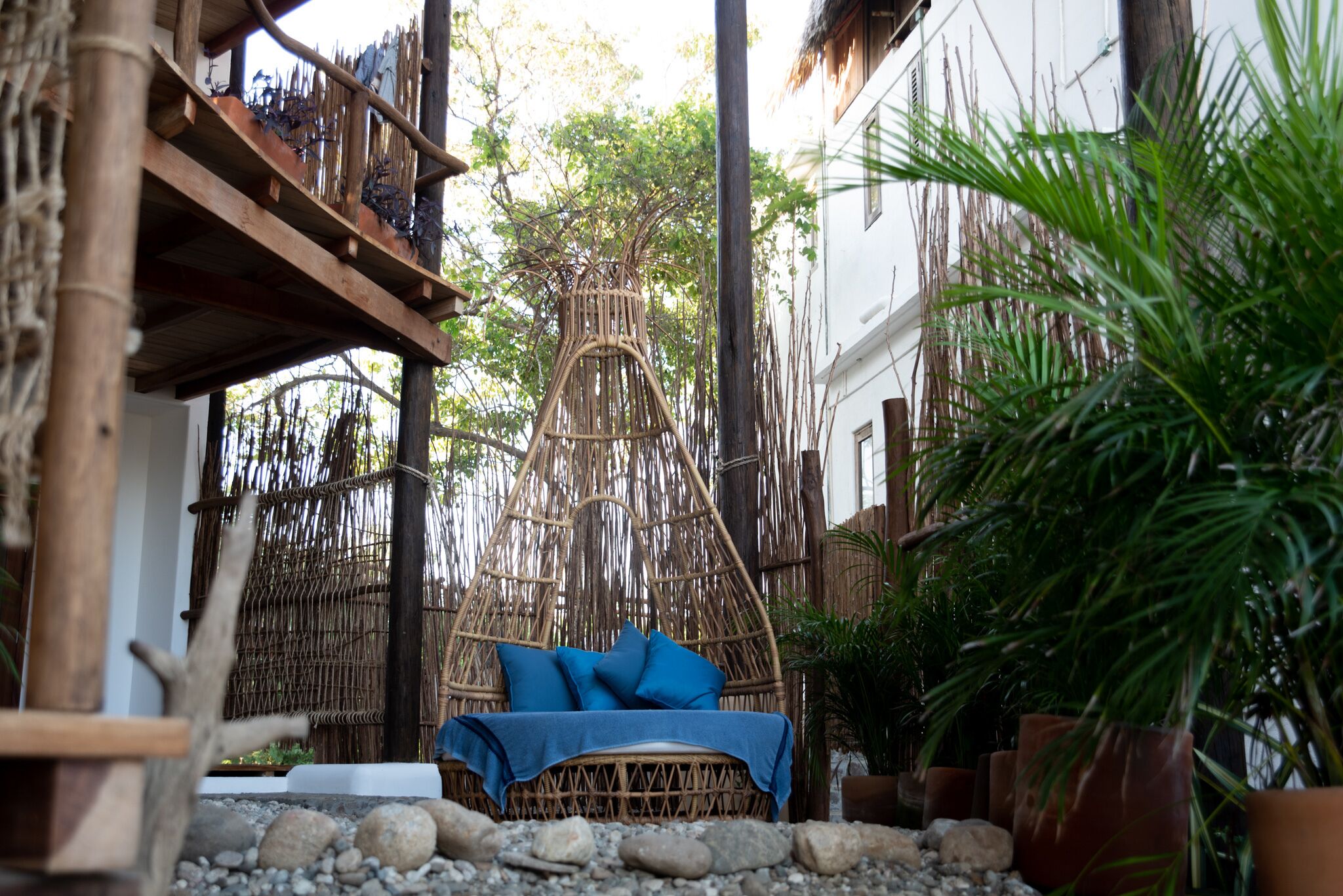 A wicker hanging chair with blue cushions in a garden area with potted plants and a bamboo fence.