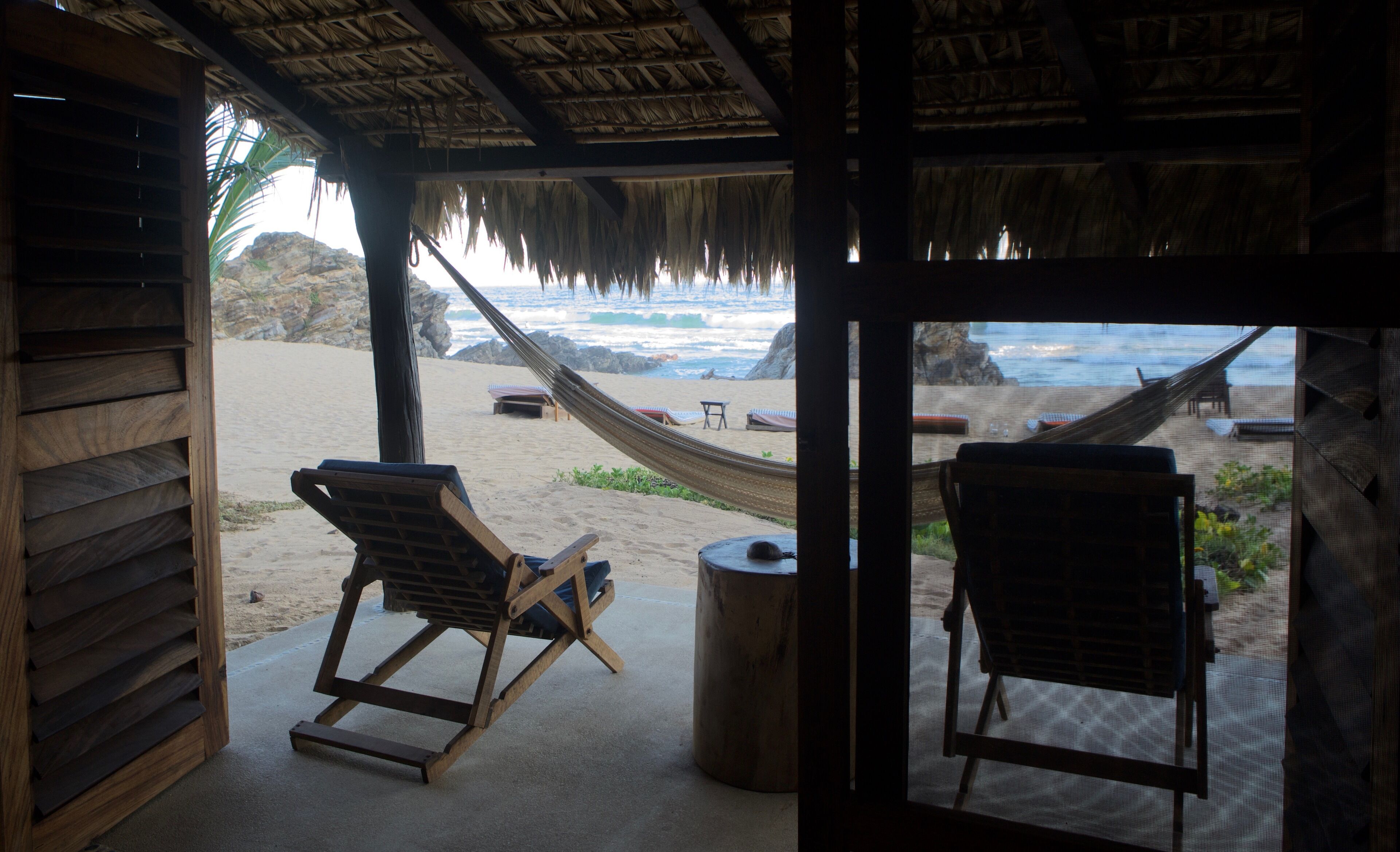A beach view from a wooden structure with a thatched roof, two wooden lounge chairs, and a hammock.