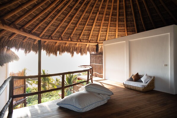 Una habitación con techo de paja, suelo de madera, una zona de estar con cojines y vistas a palmeras.