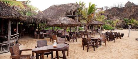 Un restaurante frente al mar con mesas y sillas de madera, sombrillas de paja y palmeras.