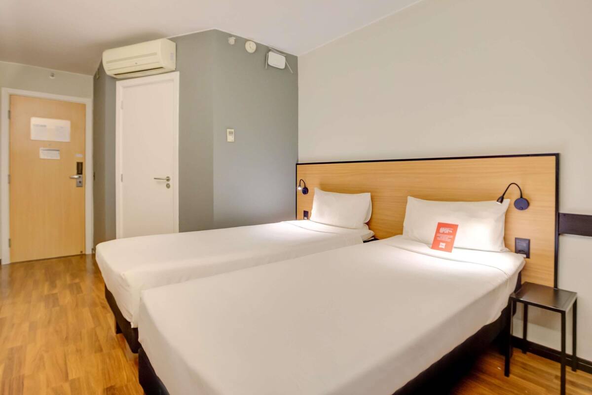Quarto de hotel com duas camas, piso de madeira, uma porta e aparelho de ar condicionado.