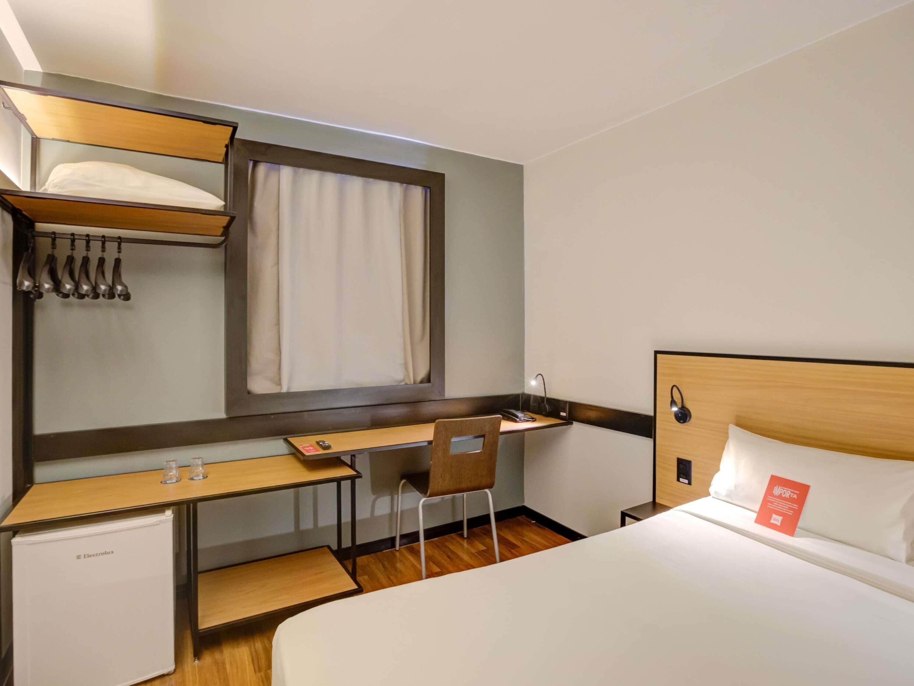 A hotel room with a bed, a desk, a chair, a small refrigerator, a window with curtains, and a wall-mounted shelf with a lamp.