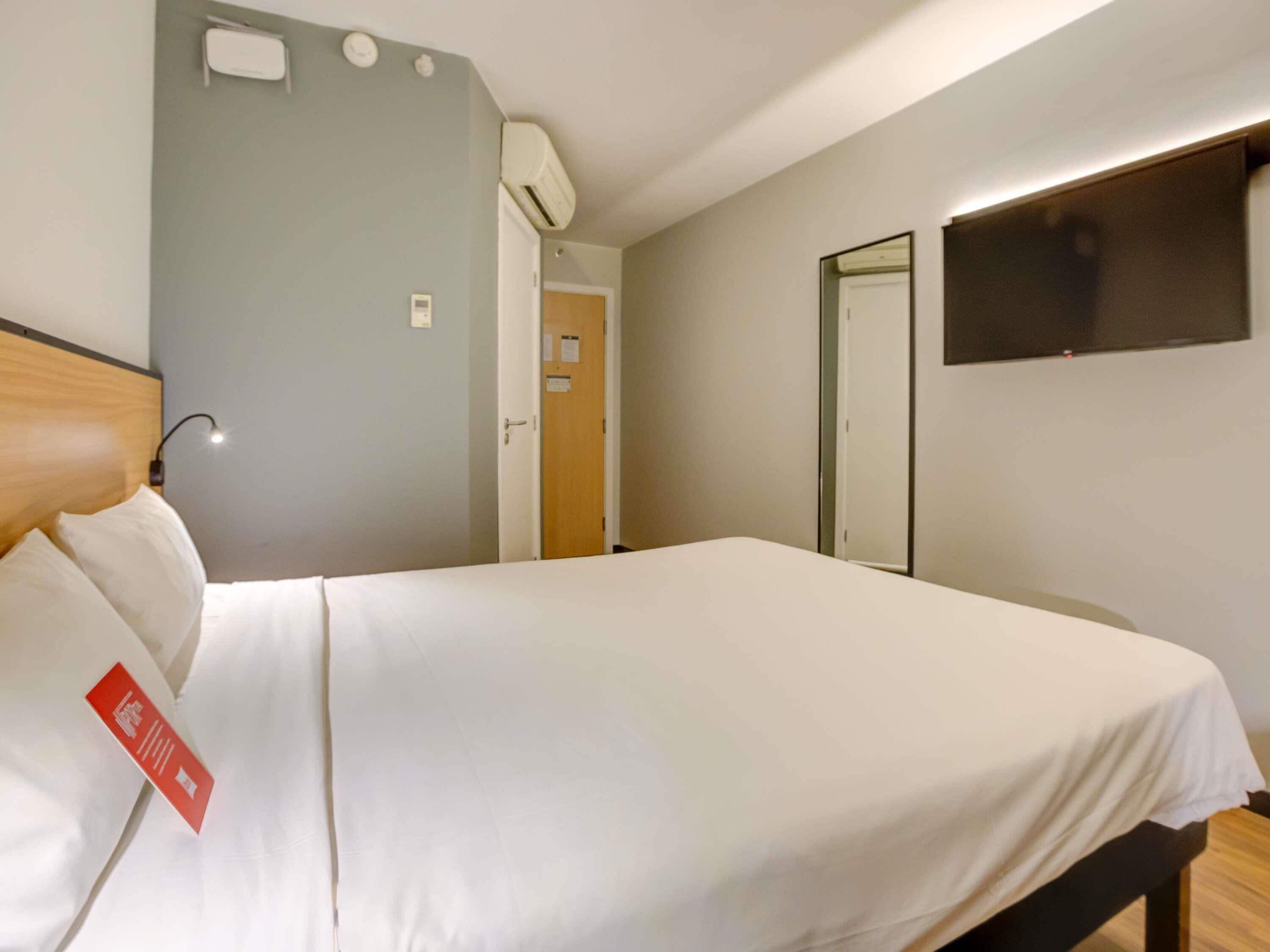 A hotel room with a bed, a bedside lamp, a wall-mounted air conditioner, and a door leading to another room.