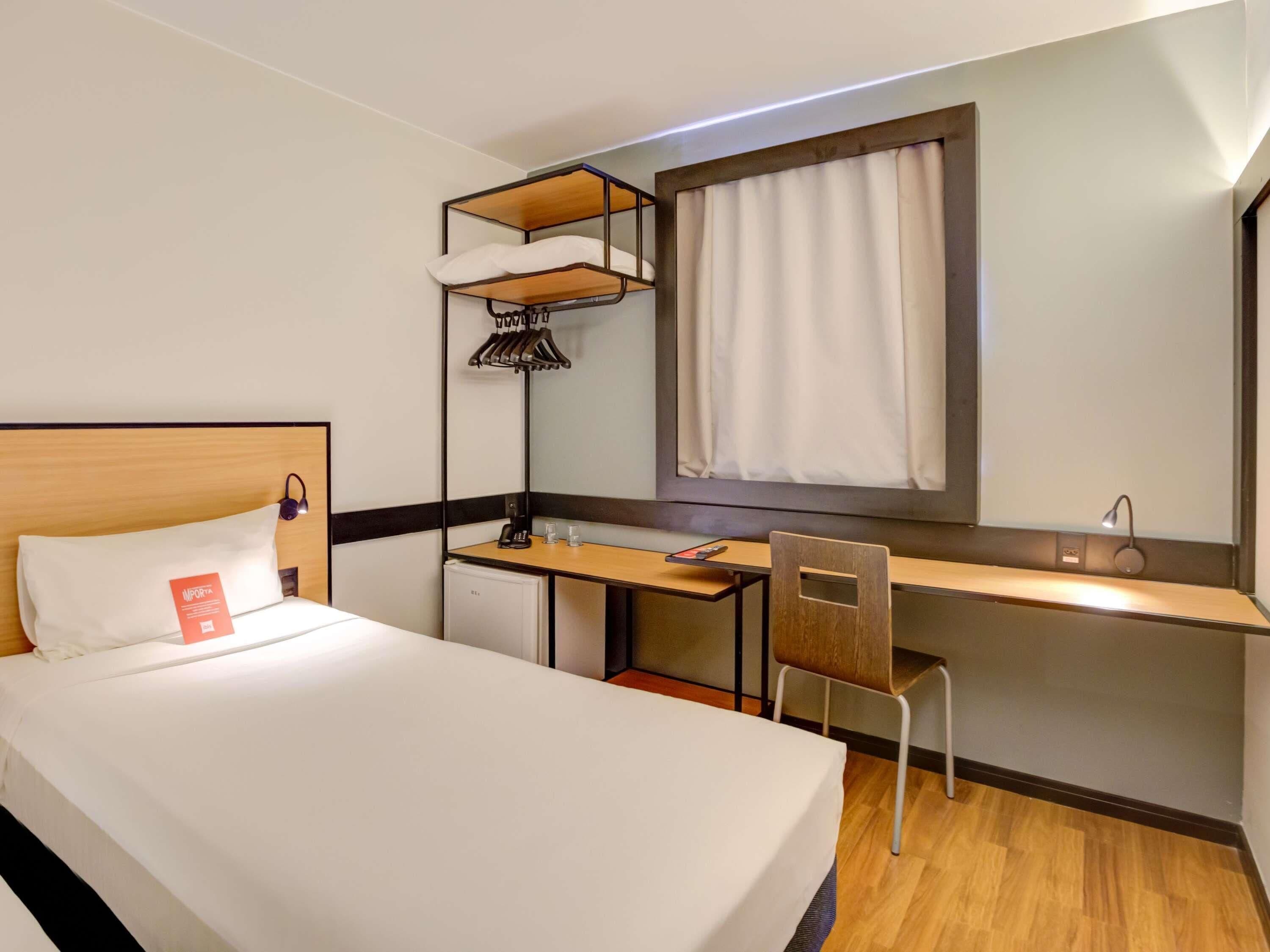 A hotel room with a bed, a desk, a chair, a small refrigerator, a hanging light, and a window with curtains.