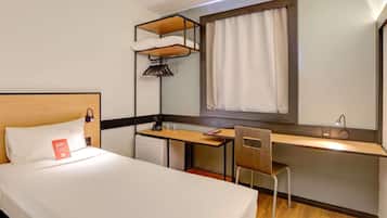 A hotel room with a bed, a desk, a chair, a small refrigerator, a hanging light, and a window with curtains.