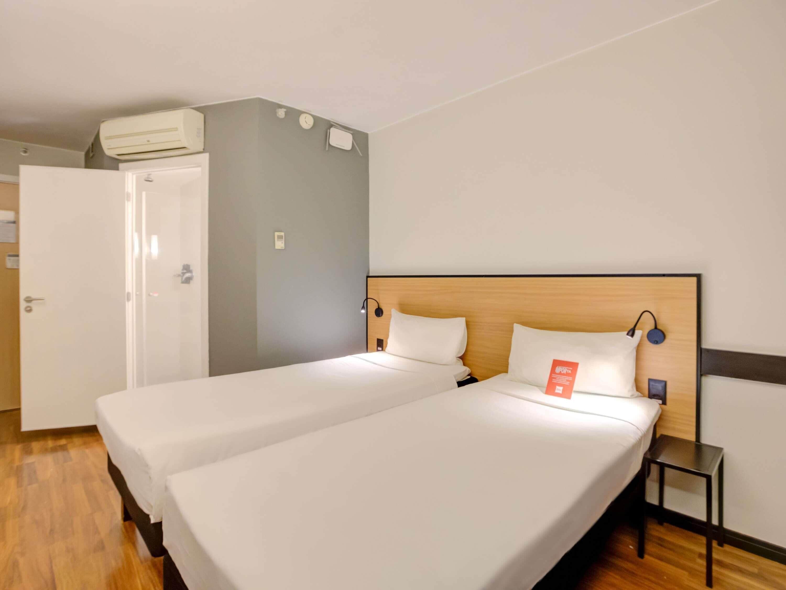 A hotel room with two beds, a wardrobe, a small table, and a wall-mounted air conditioner.