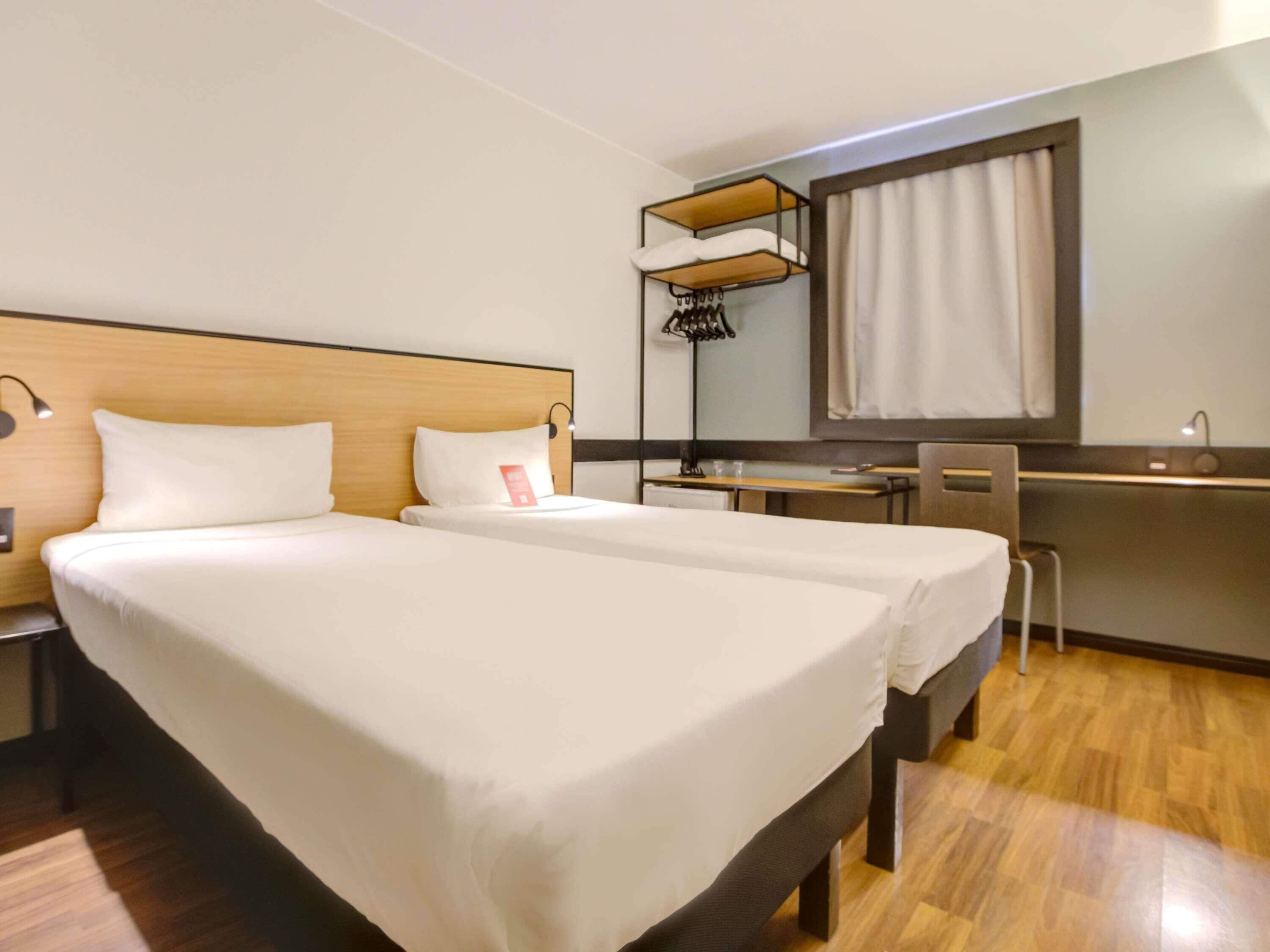 Une chambre d’hôtel avec deux lits, un bureau, une chaise, une armoire et une fenêtre avec des rideaux.