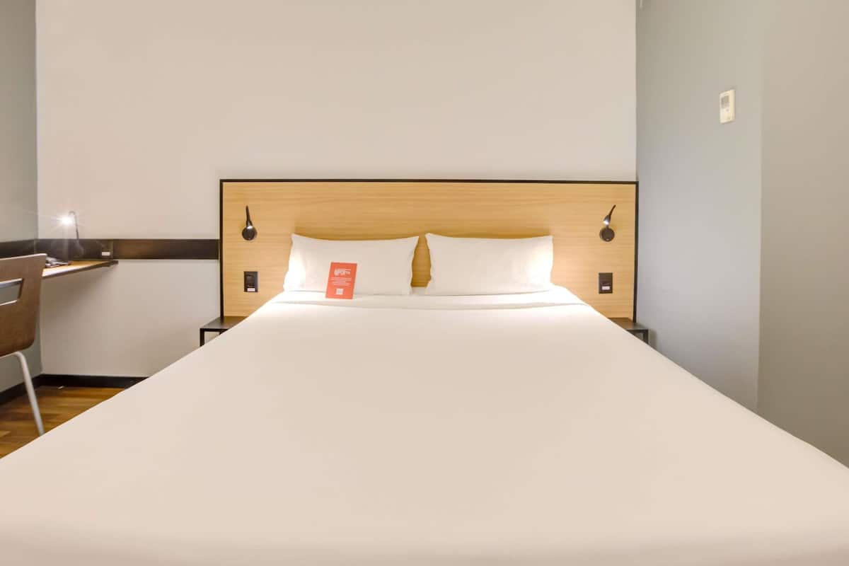 Quarto de hotel com uma cama grande, cabeceira de madeira, duas mesas de cabeceira, uma escrivaninha com cadeira e um criado-mudo.