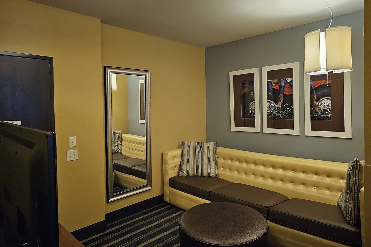 A hotel room with a large mirror, a sofa, a round ottoman, and two framed pictures on the wall.