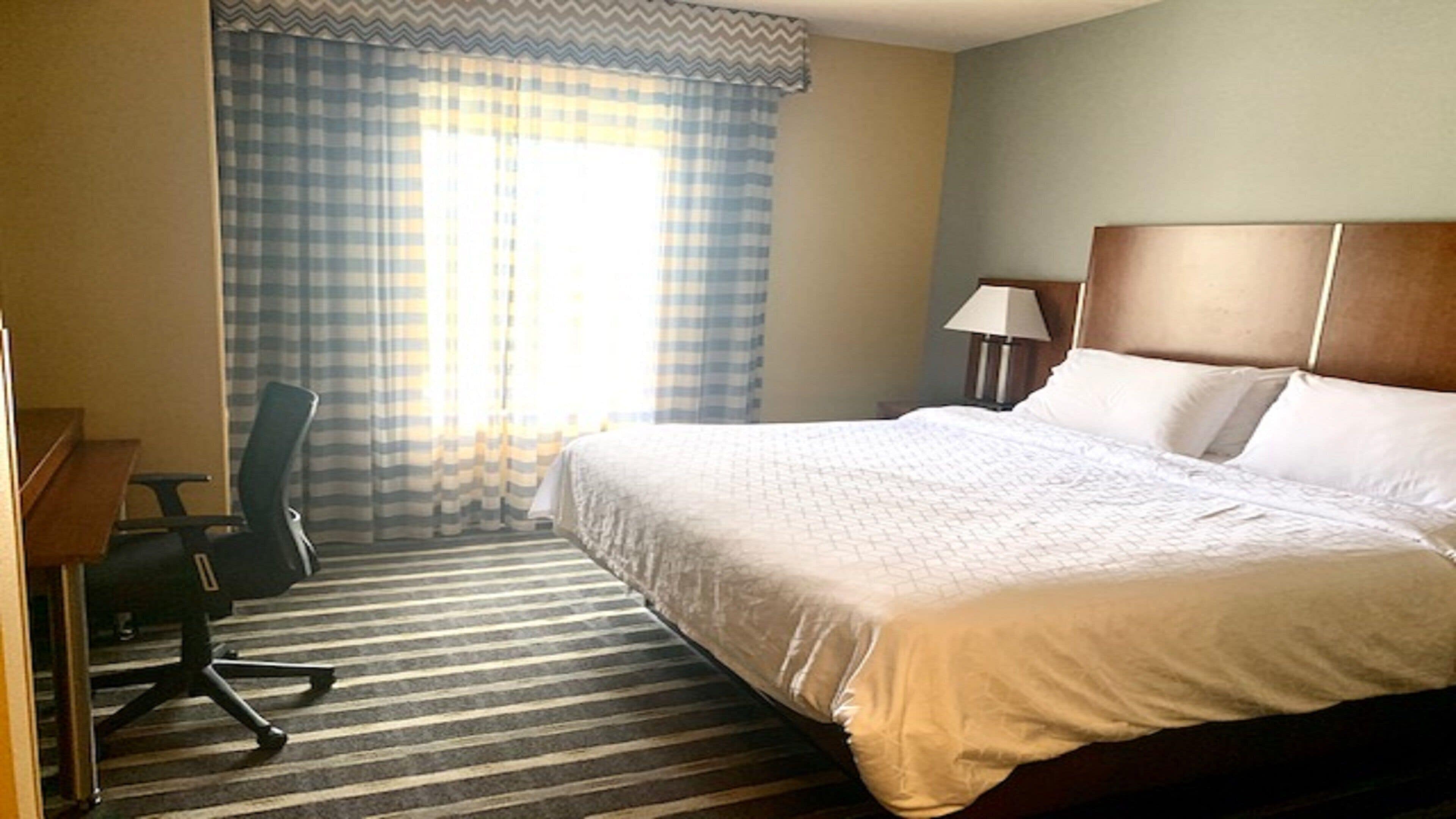 A hotel room with a large bed, a desk, a chair, a lamp, and a window with curtains.