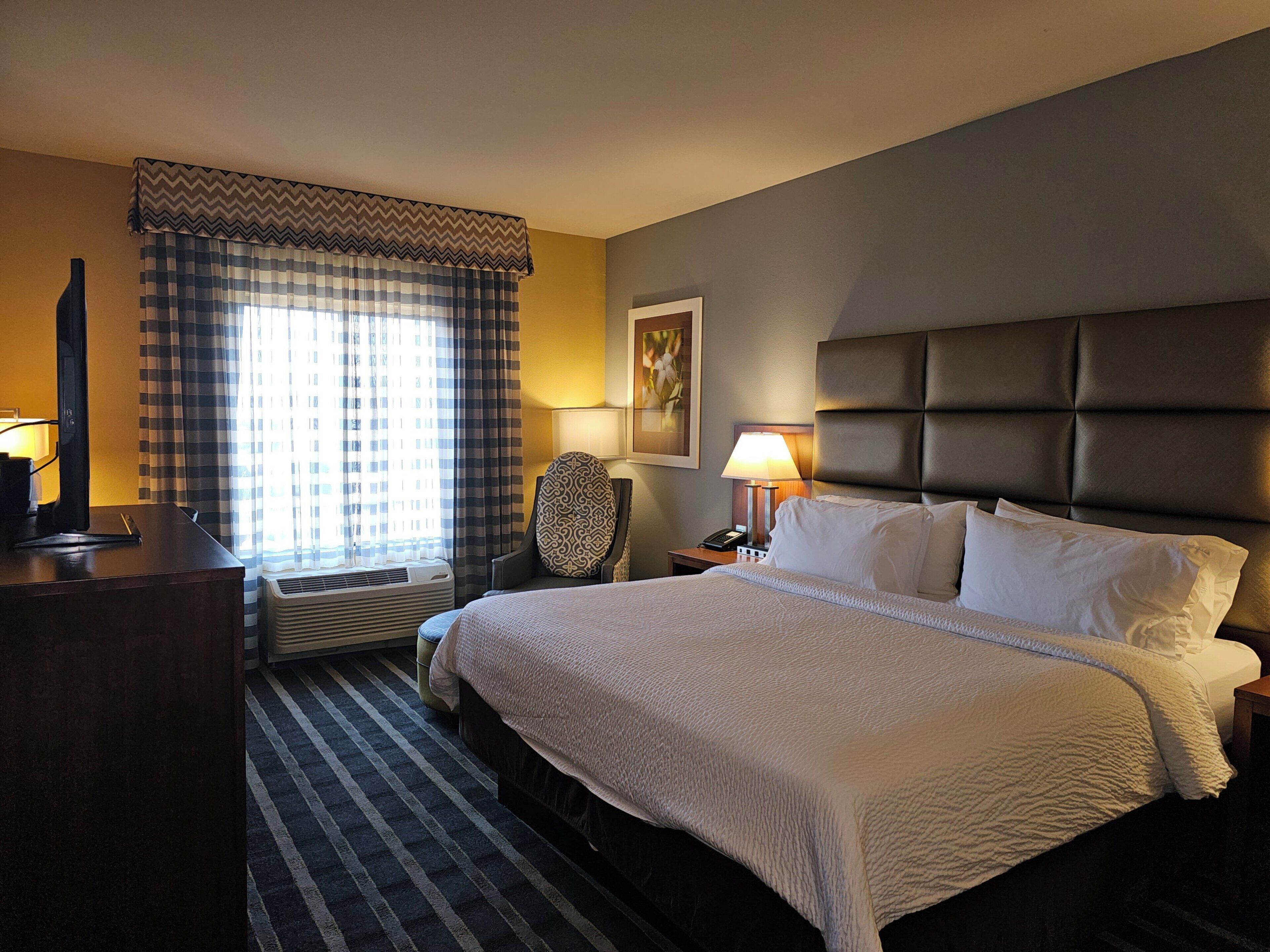 A hotel room with a bed, a chair, a desk, a lamp, and a window with curtains.