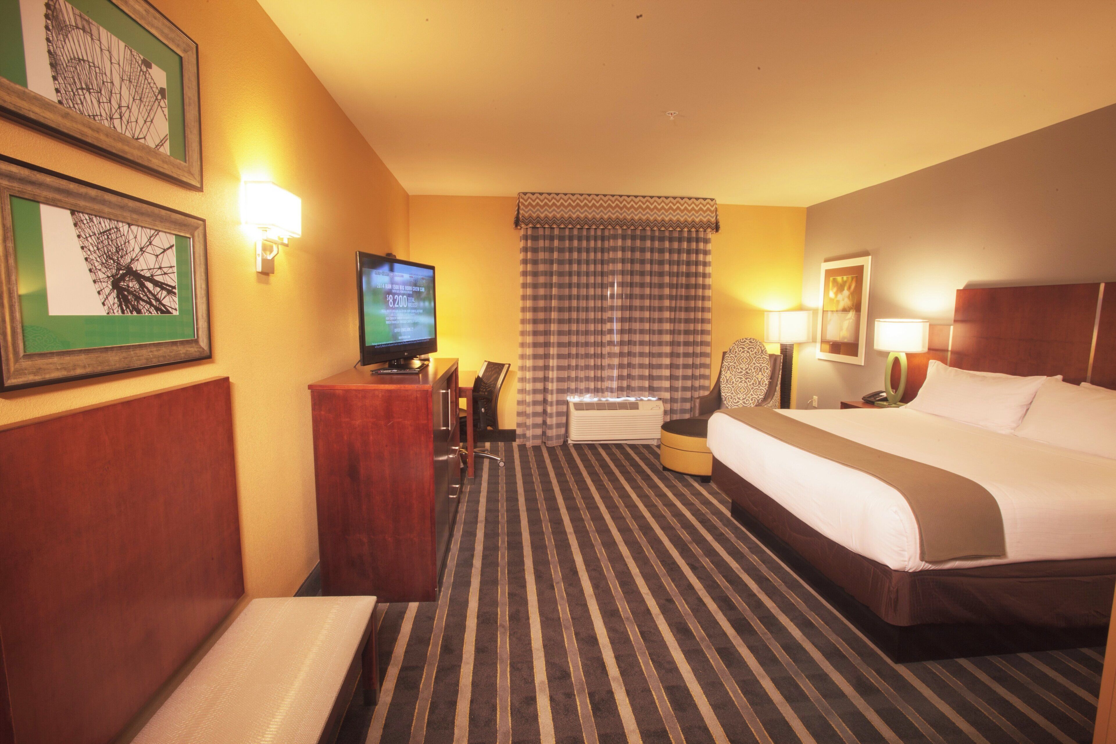 A hotel room with a bed, a desk, a television, a chair, a window with curtains, and a wall-mounted lamp.