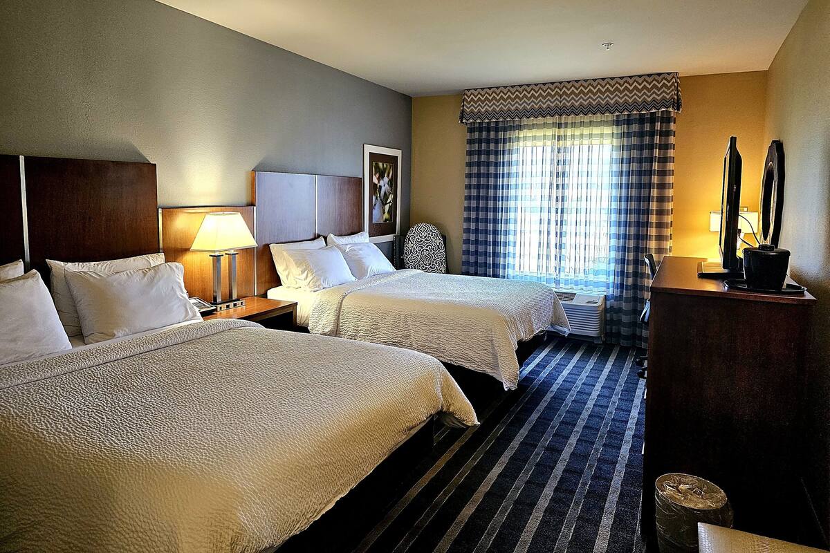 A hotel room with two beds, a desk, a chair, a lamp, and a window with curtains.