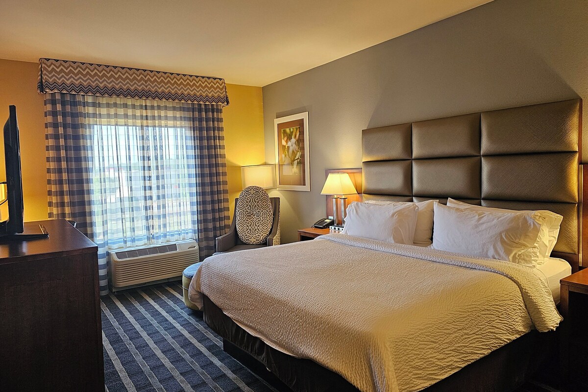 A hotel room with a large bed, a desk, a chair, a lamp, and a window with curtains.