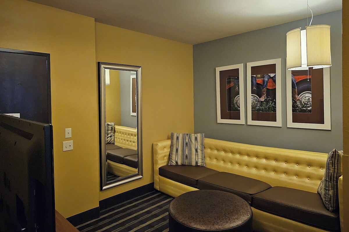 A hotel room with a large mirror, a sofa, a round ottoman, and two framed pictures on the wall.