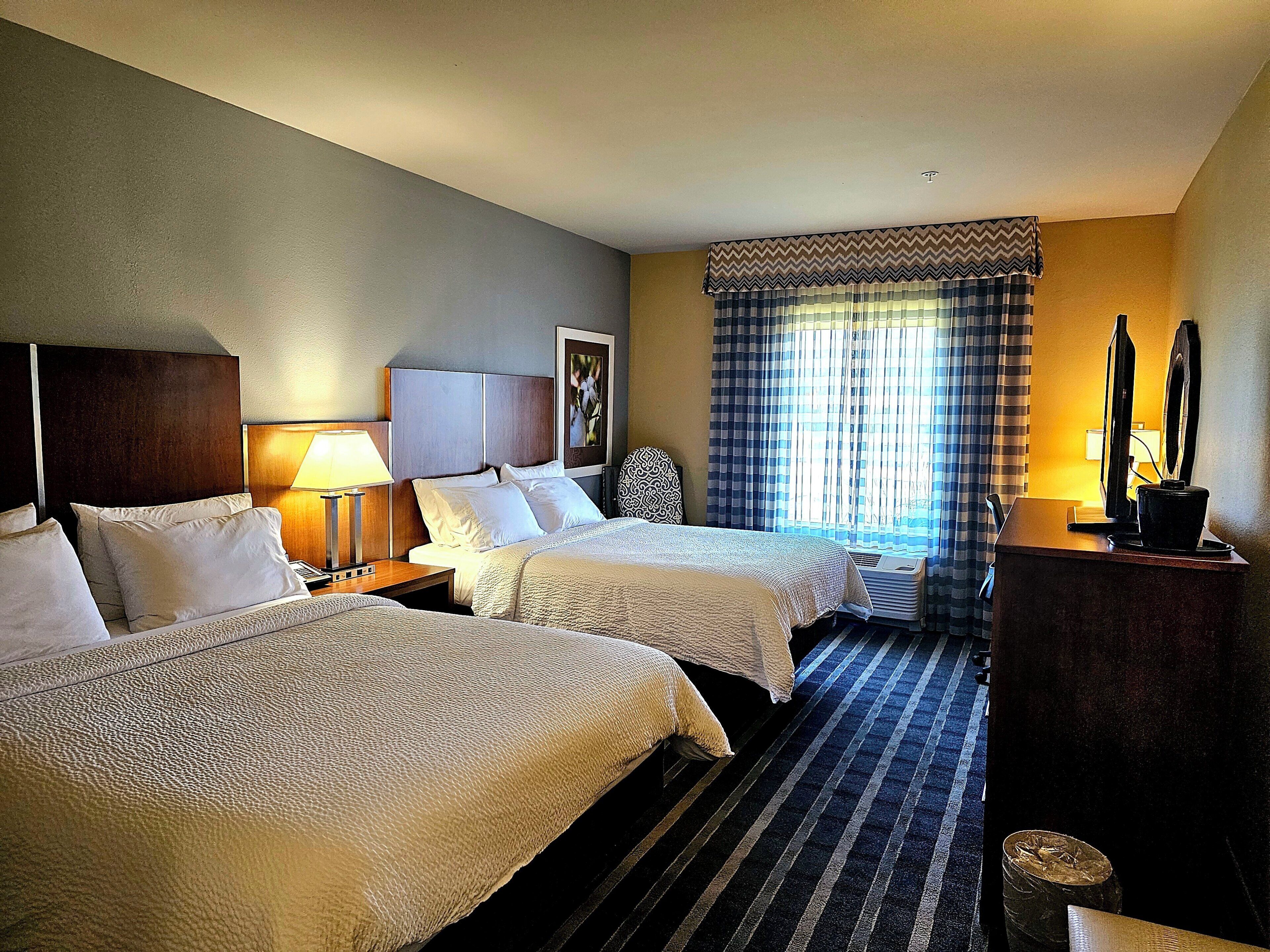 A hotel room with two beds, a desk, a chair, a lamp, a television, and a window with curtains.