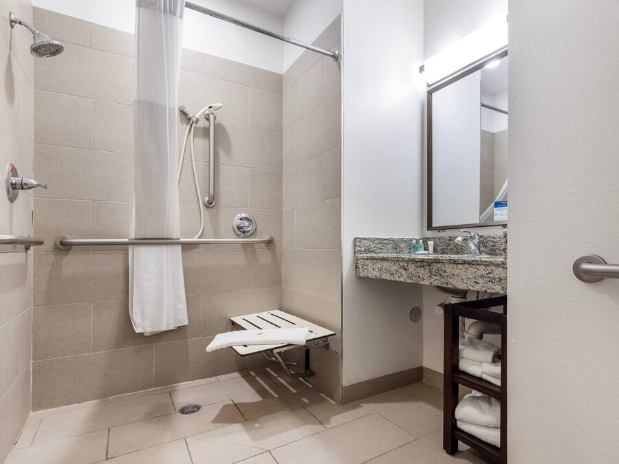 Un baño con ducha, barras de apoyo, toallero, lavabo con encimera de granito y espejo.