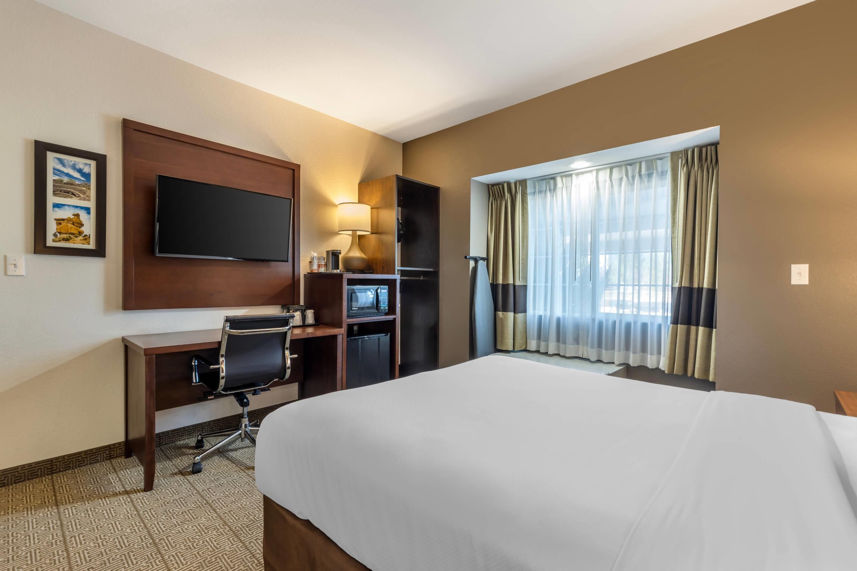 A hotel room with a large bed, a desk with a chair, a television, a desk lamp, a microwave, and a window with curtains.