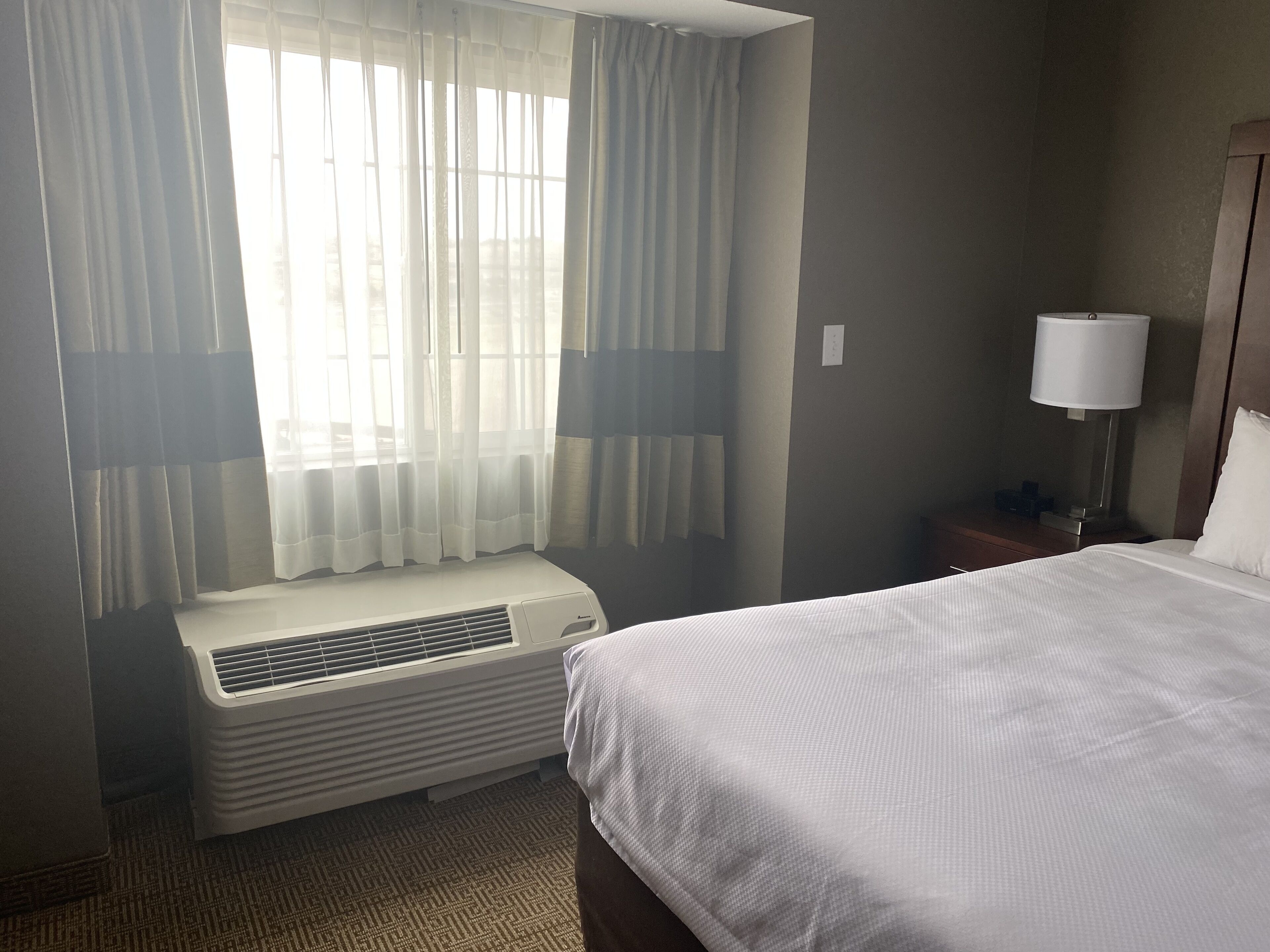 A hotel room with a bed, a nightstand with a lamp, a window with curtains, and an air conditioning unit.