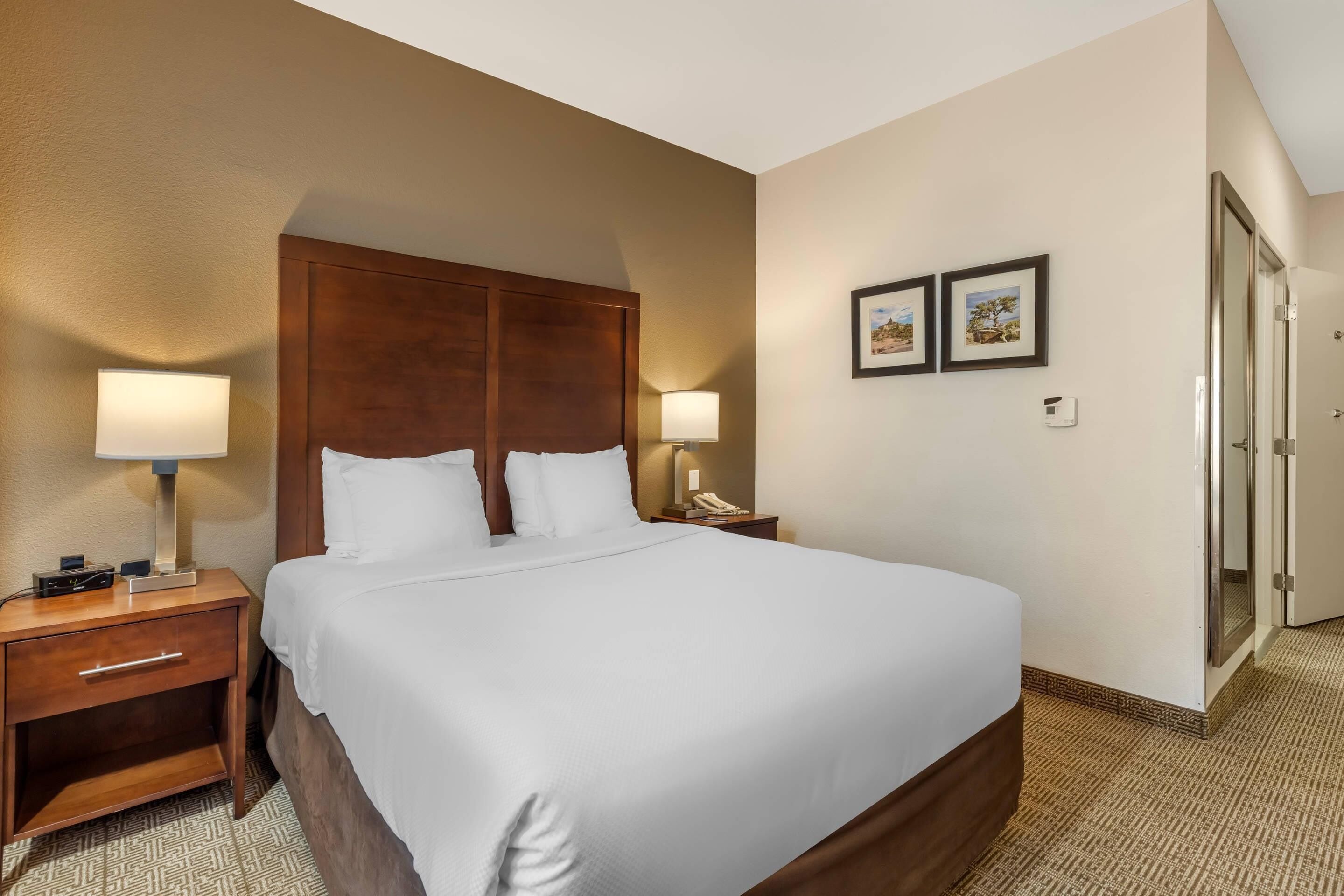 Une chambre d’hôtel avec un grand lit, une tête de lit en bois, deux lampes de chevet, une table de chevet avec un tiroir et une porte donnant sur une salle de bain.