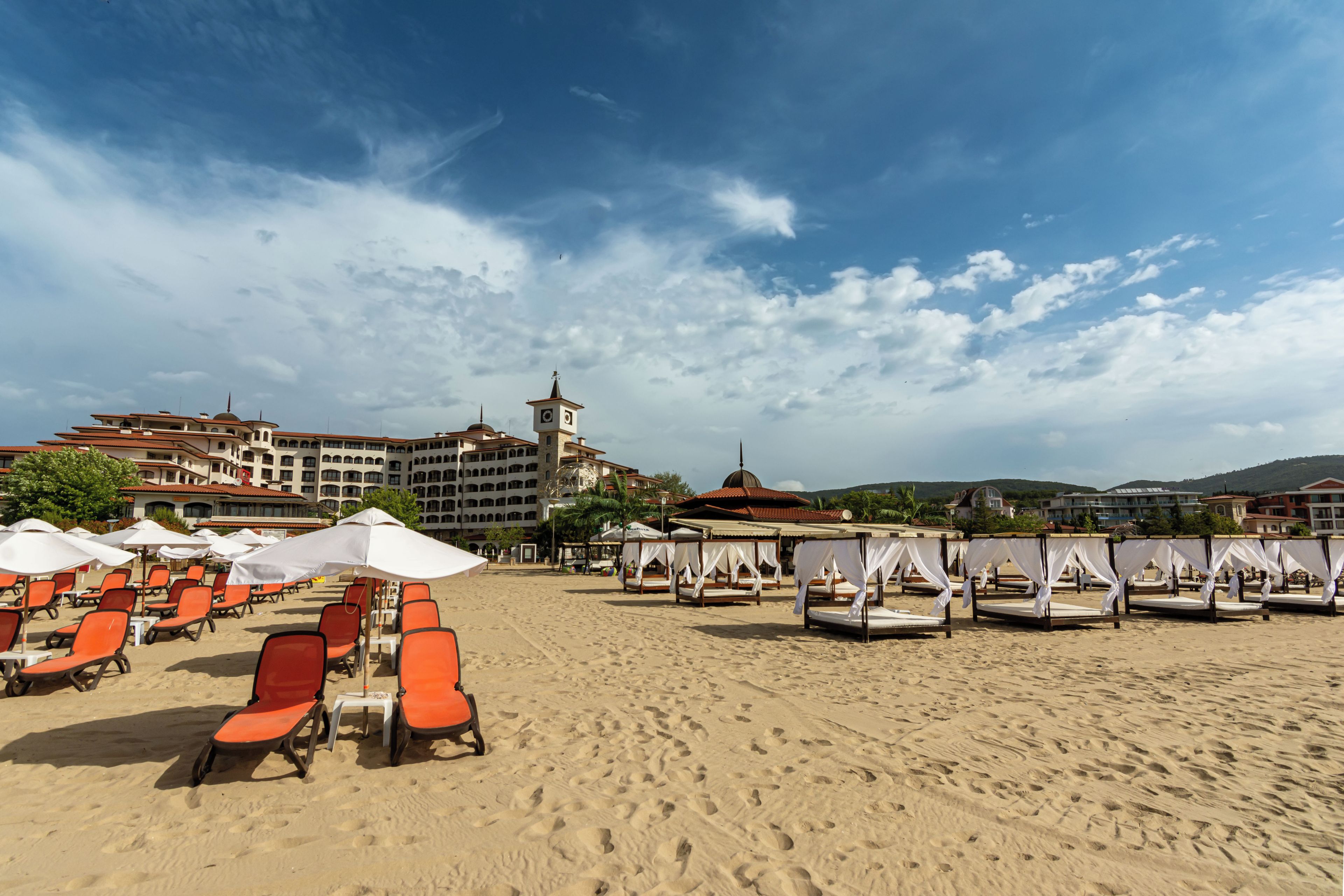 A beach with rows of orange lounge chairs and white umbrellas, a building with a clock tower, and a clear sky.