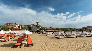 A beach with rows of orange lounge chairs and white umbrellas, a building with a clock tower, and a clear sky.