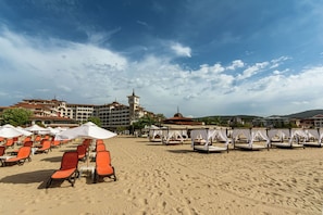 A beach with rows of orange lounge chairs and white umbrellas, a building with a clock tower, and a clear sky. - Helena Park - Ultra All inclusive (Sunny Beach)
