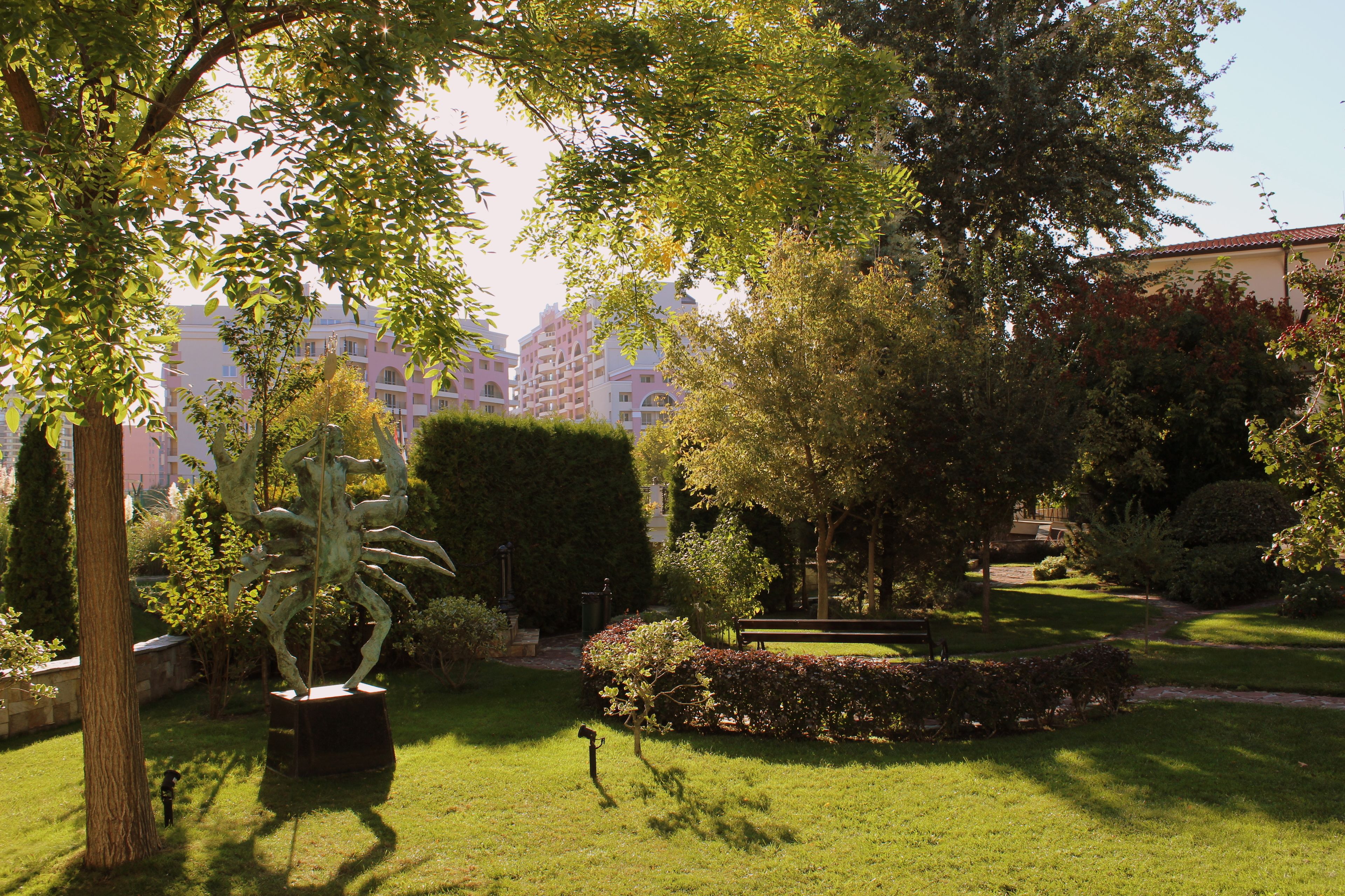 A well-maintained garden with a variety of trees and shrubs, a central sculpture, and a bench.