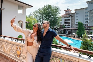 A couple taking a selfie on a balcony overlooking a swimming pool, with a view of a building with a clock tower and a tiled pool area. - Helena Park - Ultra All inclusive (Sunny Beach)