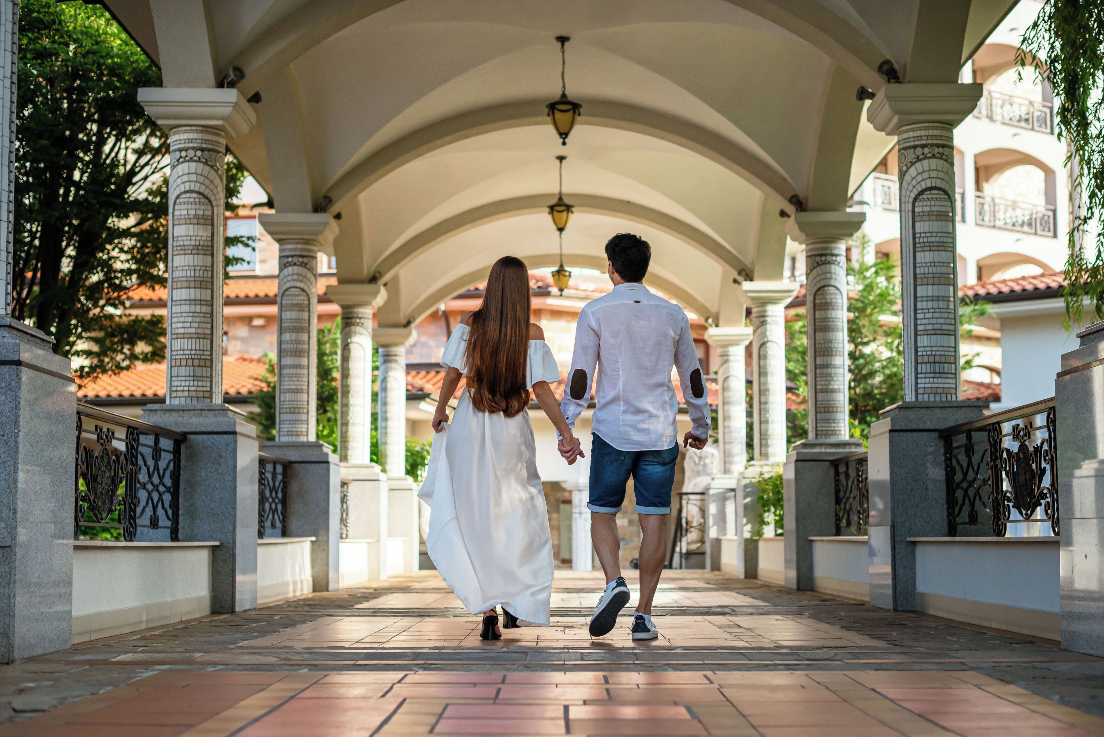 A couple walking on a tiled pathway under an arched structure with columns and railings.