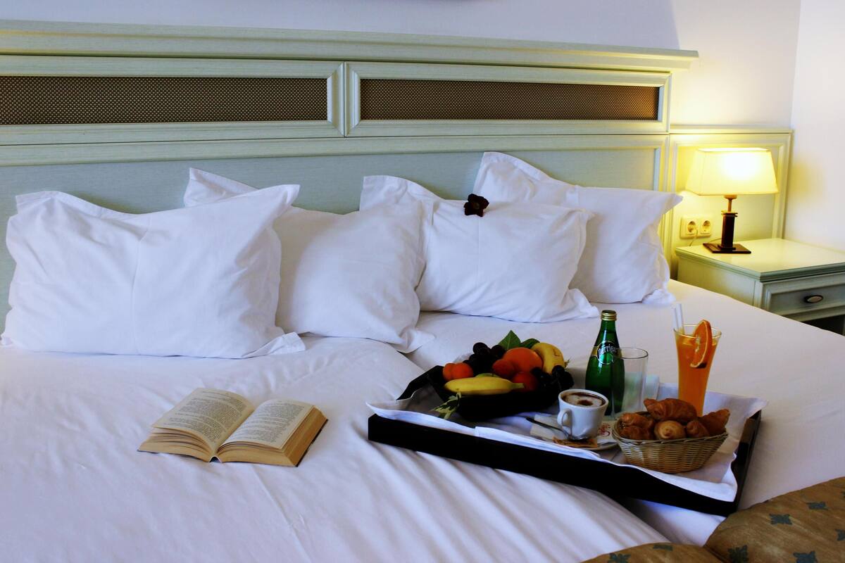 A neatly made bed with white linens, a tray with fruits, a bottle, a cup, and a basket of croissants.