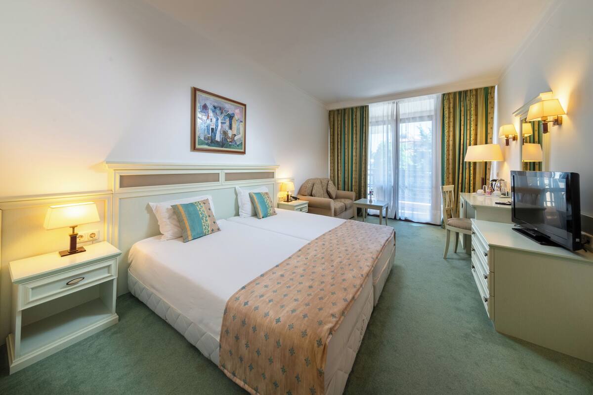 A hotel room with a large bed, a desk with a television, a chair, and a desk lamp.