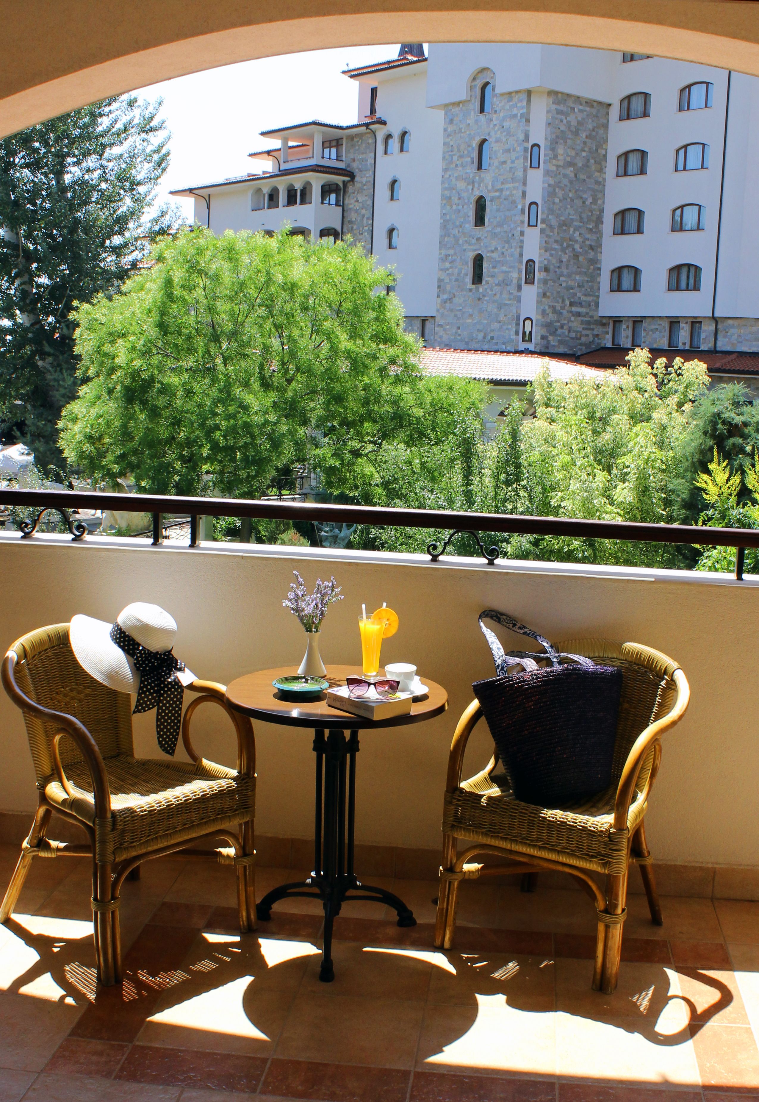 Ein Balkon mit Korbstühlen, einem Tisch mit einer Vase und einem Getränk, und Blick auf ein Gebäude und Bäume.