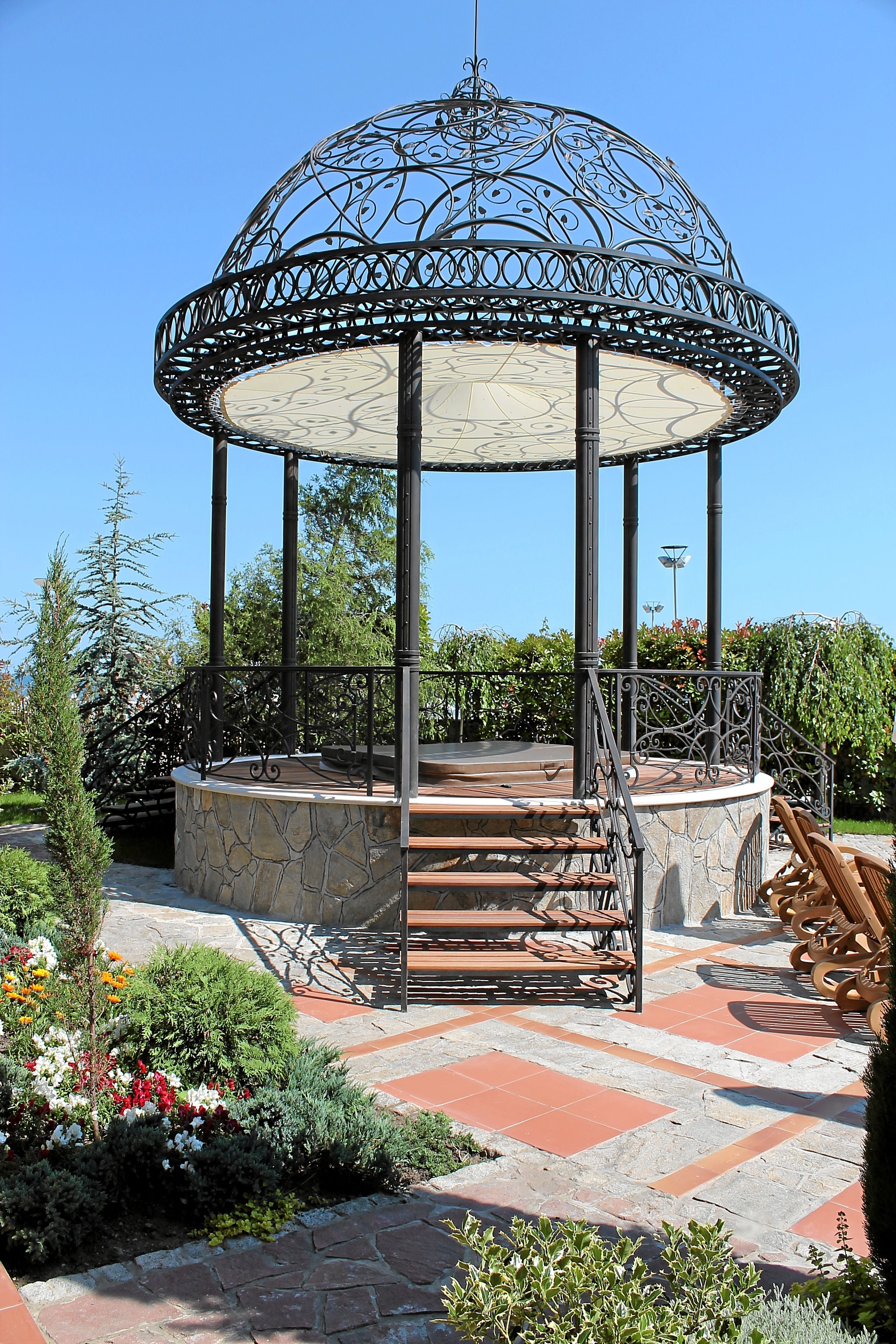 A gazebo with a stone base and metal railing, surrounded by a garden with various plants and flowers.