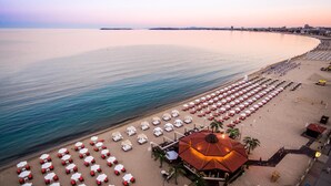 A beach with rows of sunbeds and umbrellas, a central building with a red roof, and a calm sea. - Helena VIP Villas and Suites (Sunny Beach)