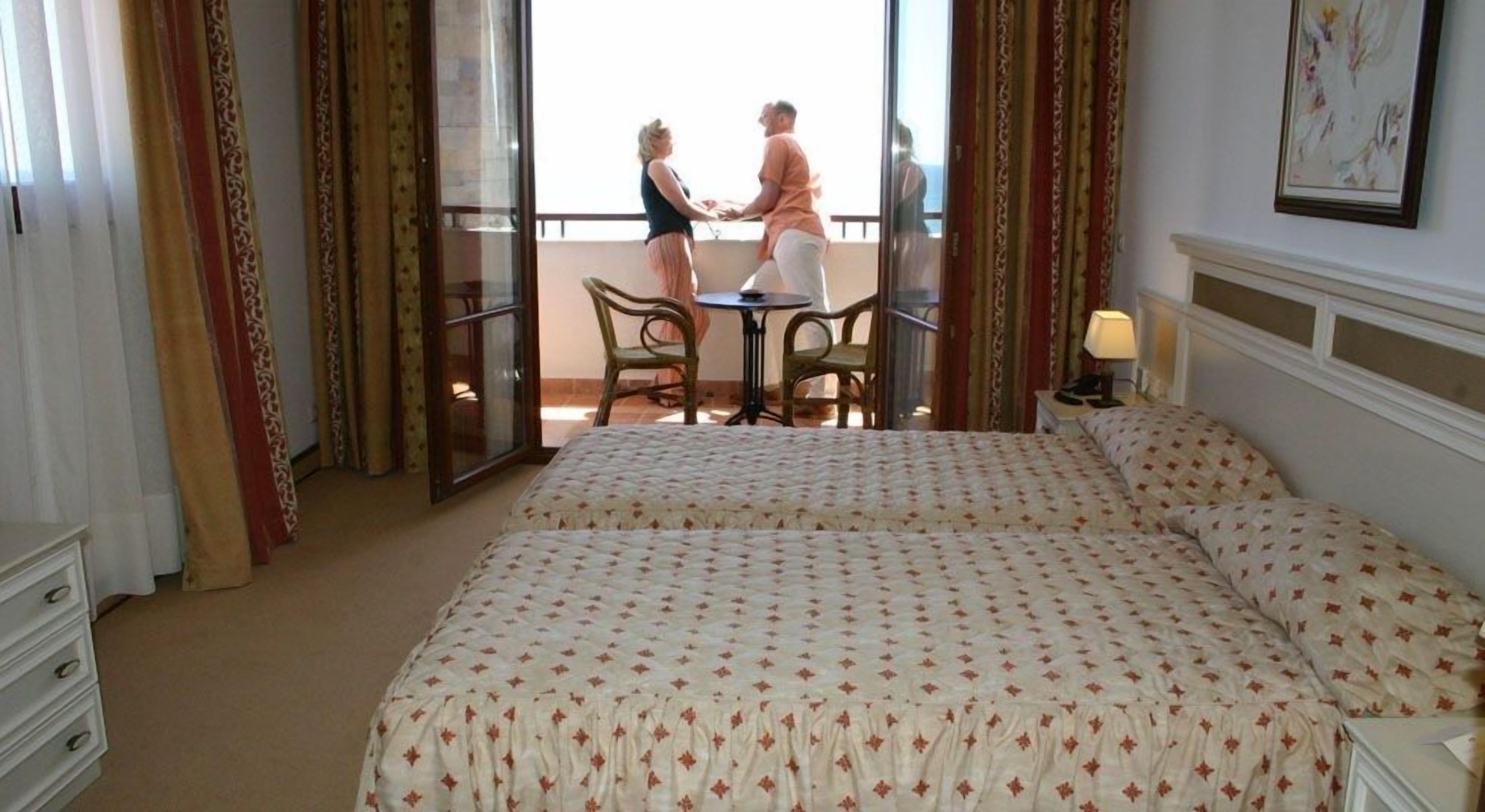 A hotel room with two beds, a balcony with a table and chairs, and a view of the sea.