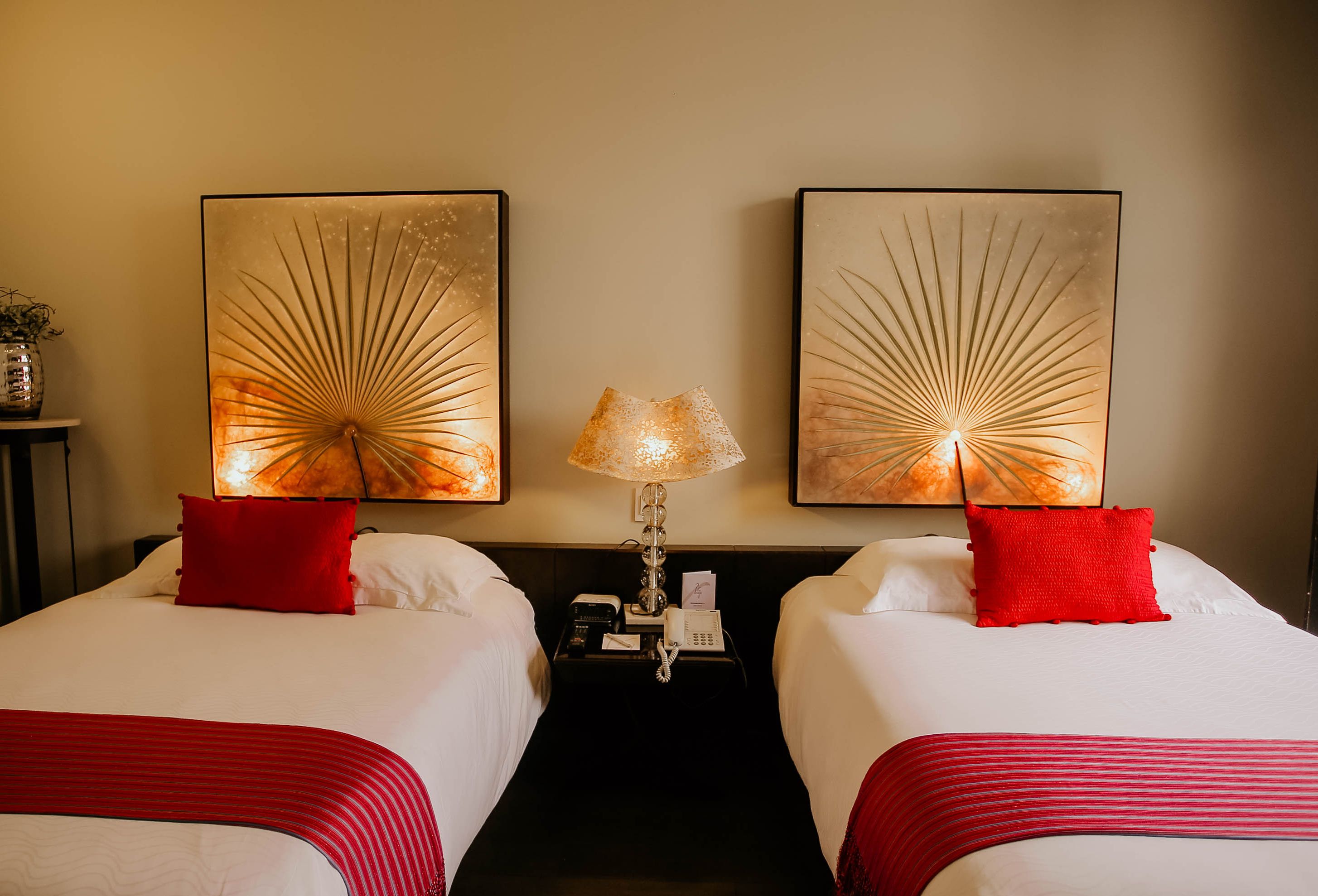 A hotel room with two beds, each with a red pillow and a red-striped bedspread. There are two wall-mounted lamps with a sunburst design, and a nightstand with a telephone and a small table with a vase of flowers.