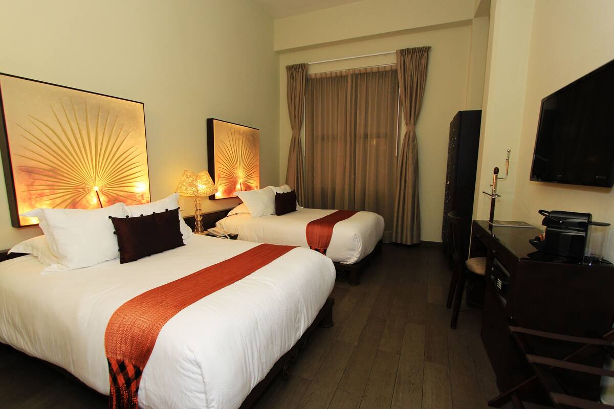 Quarto de hotel com duas camas, uma escrivaninha, televisão e obras de arte nas paredes.